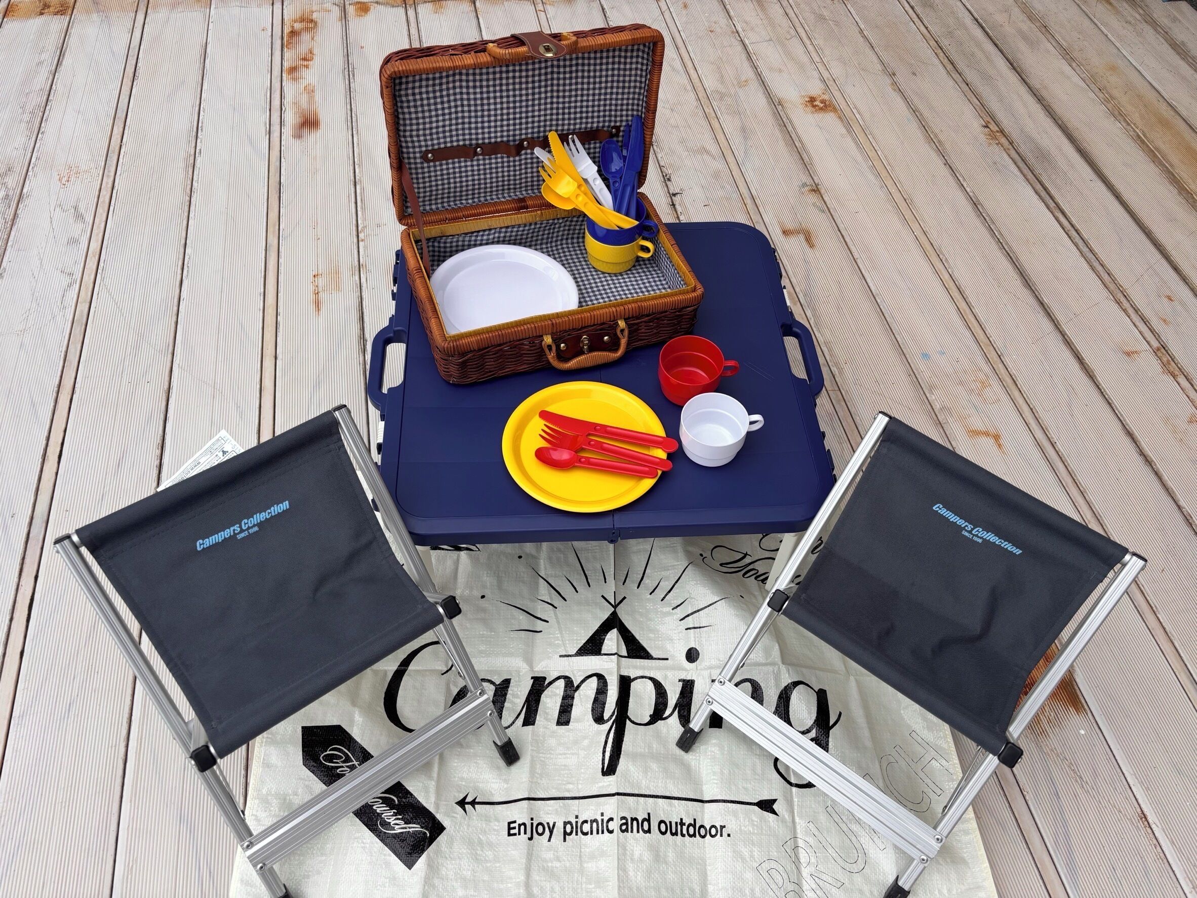 A picnic setup with a basket, utensils, a plate, and a cup on a wooden deck.