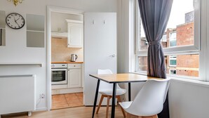 A compact kitchen with white cabinets and a built-in oven, a dining area with a table and chairs, and a window with curtains. - Blueground | Lambeth, wifi, nr Kennington Park (London)