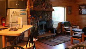 A rustic cabin interior with a stone fireplace, wooden furniture, and a kitchen area. - Cozy Private Lakefront Terry Redlin-Style Cabin w/ Unique Loft (Brainerd)