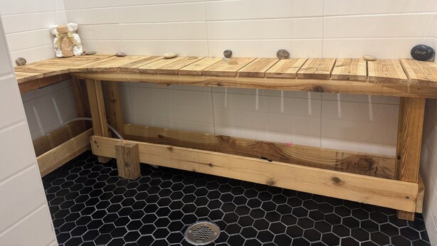 A corner bathroom with a wooden bench, a small white teddy bear, and a hexagonal black floor.