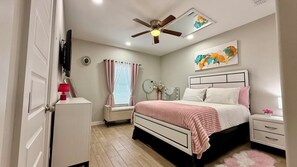 Un dormitorio con cama, mesitas de noche, ventilador de techo, ventana con cortinas y un reloj de pared.