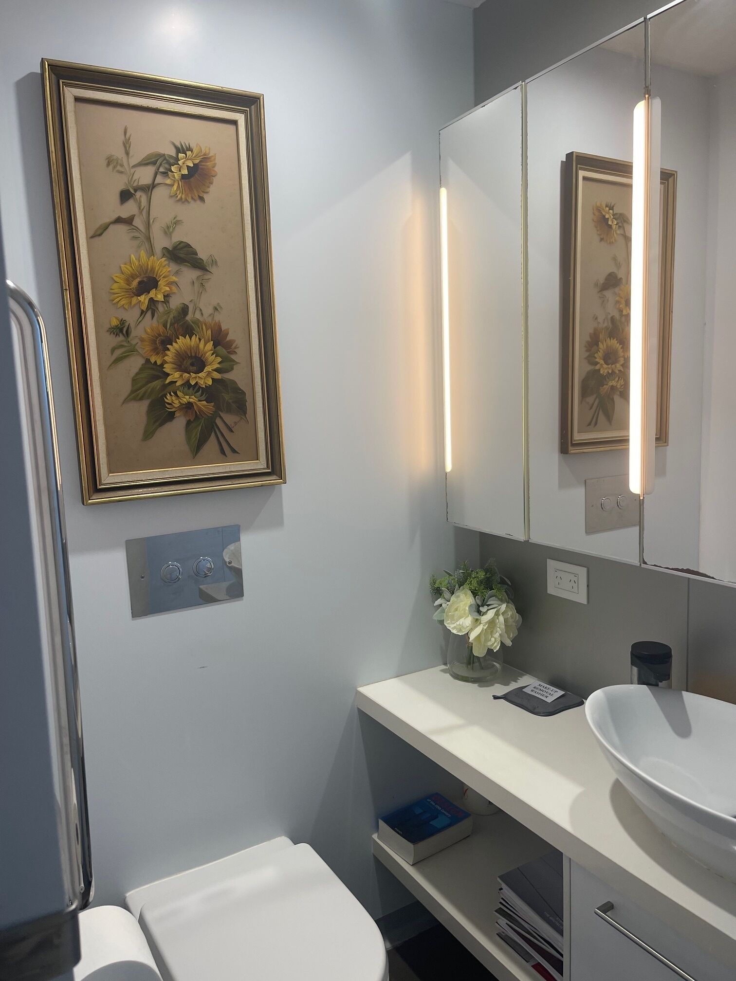 Un baño con un lavamanos blanco, un espejo y una obra de arte floral enmarcada en la pared.