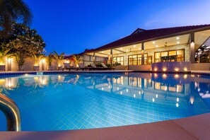 A swimming pool with a tiled deck, a covered outdoor seating area, and a stone facade building in the background. - Lovely 3BR Villa in good Location CV3MA (Hua Hin)