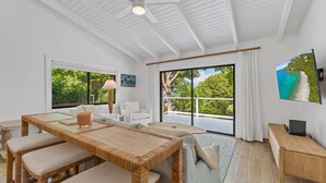 A spacious living room with a large window, a dining area with a wooden table, and a balcony with a view of the greenery. - Private Pool | 2 Mins to Beach | Walk to Dining (St. Thomas)