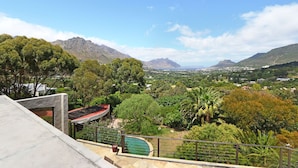 Vista de uma varanda com paisagem verdejante, montanhas ao longe, piscina e um edifício moderno com telhado vermelho.