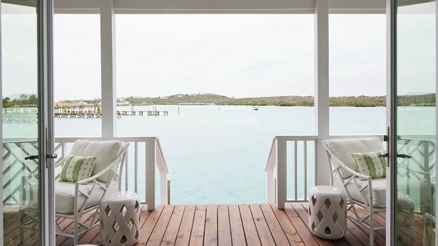 Un balcón techado con sillas blancas y una terraza de madera, con vista a una masa de agua tranquila y una costa distante.