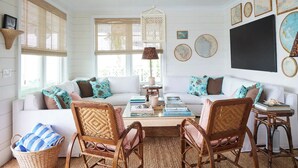 Un salón acogedor con un sofá blanco, sillas de mimbre, una mesa de centro con libros y una lámpara colgante.