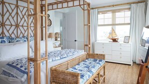 Una habitación con una cama con dosel, un banco de mimbre, un aparador blanco y un ventanal con cortinas.
