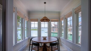 A dining area with a round table and chairs, large windows with views of a body of water and buildings, and a hanging light fixture. - South End Villa on the Water: Lovely Pool, Expansive Decks, and 60’ Private Dock (Elbow cay)
