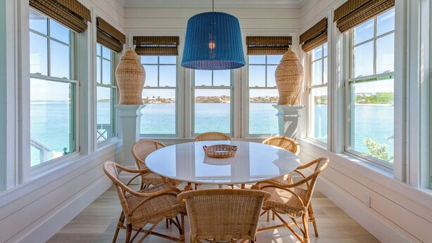 Mesa redonda de comedor con sillas de mimbre y una lámpara colgante azul en una habitación con ventanas grandes que dan a una masa de agua.