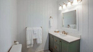 A bathroom with a white vanity, a green cabinet, a white toilet, and a mirror with three lights. - Ocean View Cottage at Abaco Inn (Elbow cay)