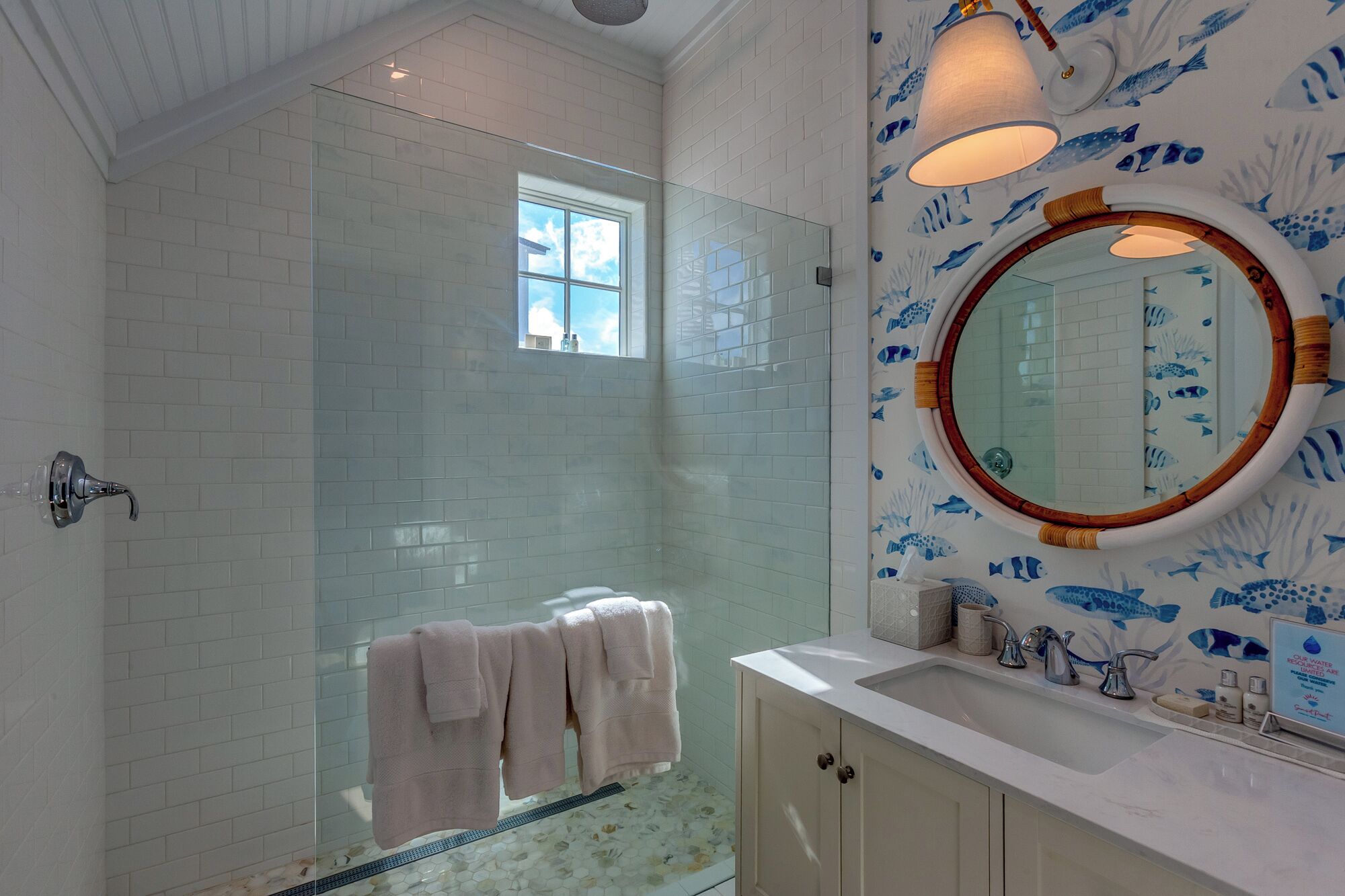 Ein Badezimmer mit Glasdusche, weißen Fliesenwänden, einem runden Spiegel mit Holzrahmen und einem Waschbecken mit weißer Arbeitsplatte.