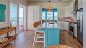 Une cuisine avec des meubles blancs, un îlot central bleu, un luminaire suspendu et une vue sur l’océan grâce à de grandes fenêtres.