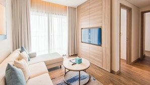 Une chambre d’hôtel moderne, dotée d’un canapé, d’une table basse ronde, d’un téléviseur à écran plat fixé au mur et d’un mur revêtu de panneaux en bois.