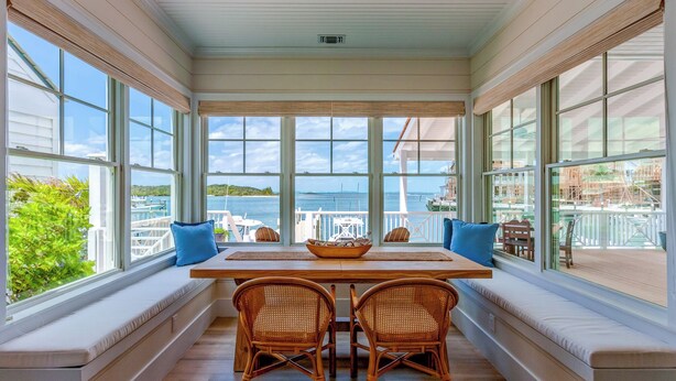 A sunlit room with a large window overlooking a body of water, a wooden table, two wicker chairs, and a bench with cushions.