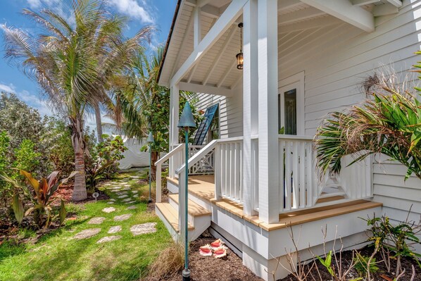 Une maison blanche avec un porche abrité, un chemin de pierre et des plantes tropicales.
