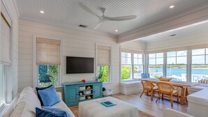 A spacious living room with a large TV, a dining area with wicker chairs, and a view of the water.
