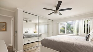 Une chambre à coucher avec un lit, un ventilateur de plafond, une fenêtre avec des stores et un miroir.