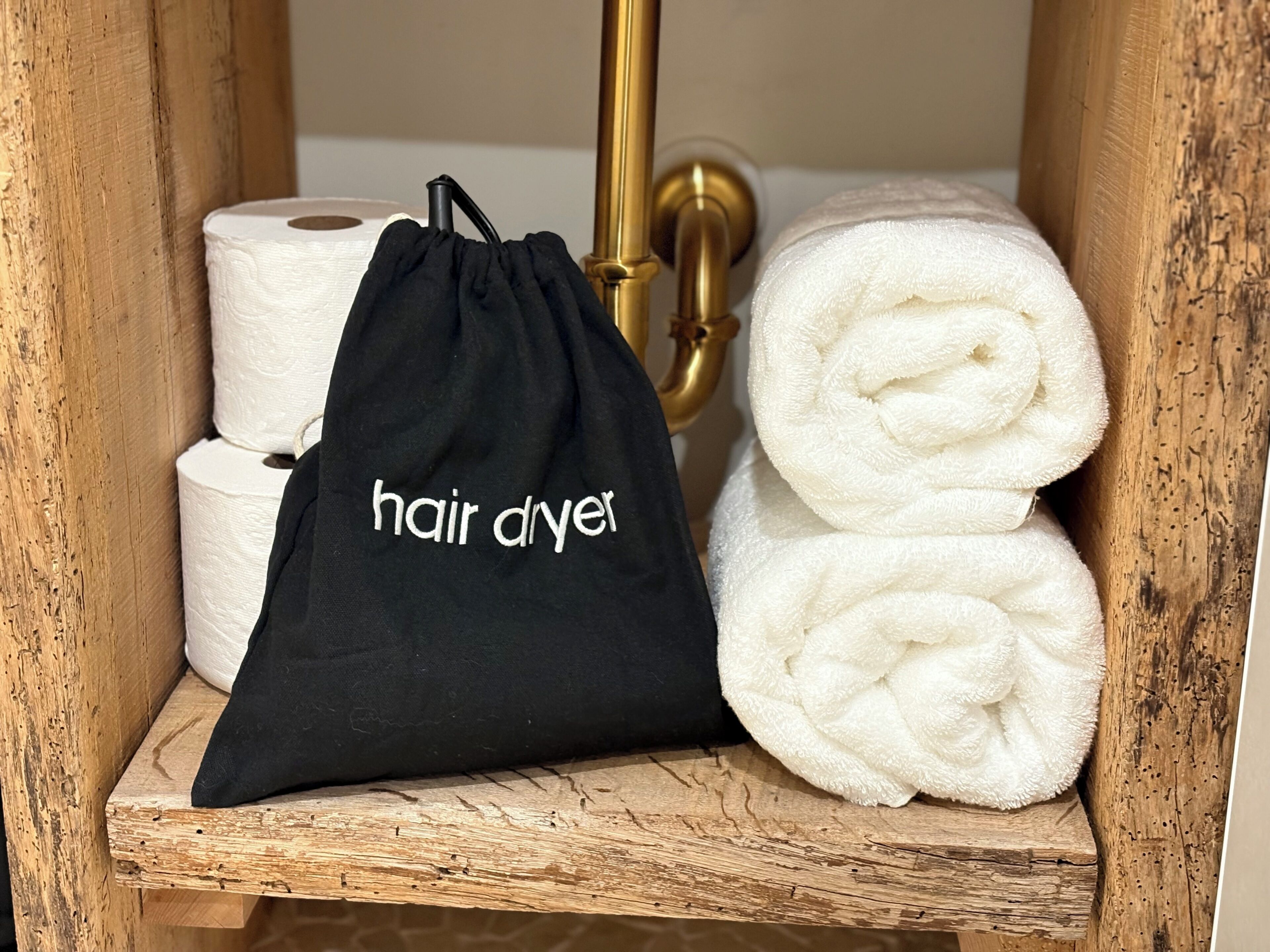 Une étagère en bois sur laquelle se trouvent un sac noir à cordon de serrage étiqueté « sèche-cheveux », des serviettes blanches et du papier toilette.