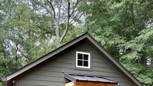 Una casa pequeña de un solo piso con techo inclinado, un porche con viga de madera y una puerta con ventana encima. A cada lado de la puerta hay dos ventanas pequeñas, y un pequeño porche con plantas en macetas.