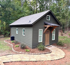Una pequeña casa de un piso con revestimiento exterior oscuro, un porche con viga de madera y un sendero de piedra que conduce a la entrada.