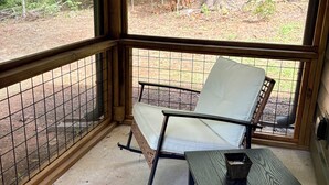 Varanda com estrutura de madeira, uma cadeira de vime e uma pequena mesa, com vista para uma área arborizada.