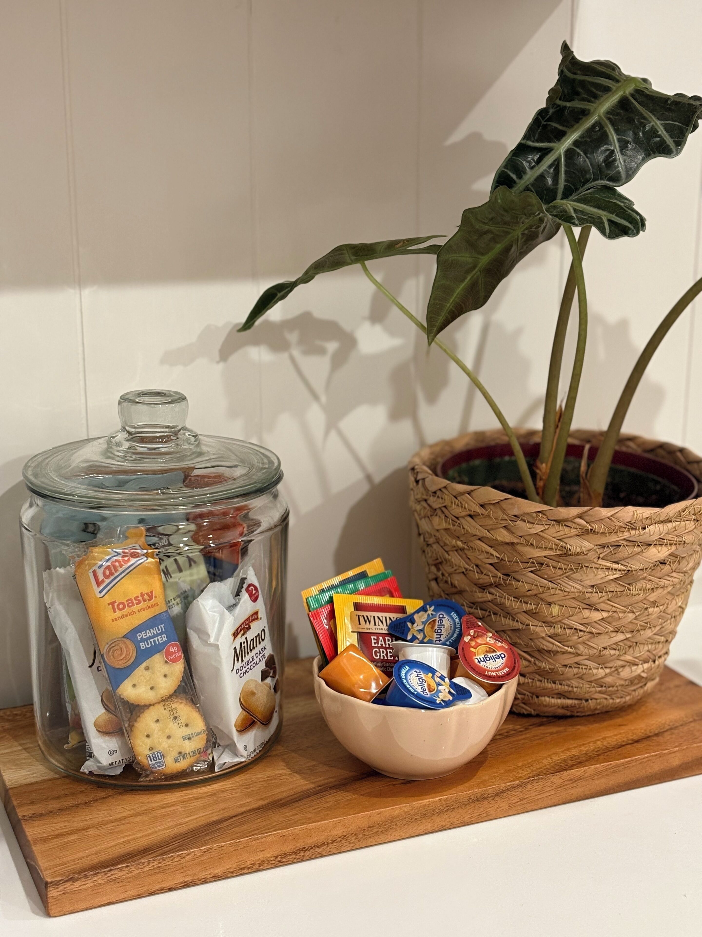 A glass jar with snacks, a bowl of assorted chocolates, and a potted plant on a wooden tray.