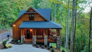 A wooden cabin with a porch, stone foundation, and a view of the forest. - Pearl's Mountain Retreat: Wifi, Hot Tub, Free Attraction Tickets, & Game Room (Sevierville)