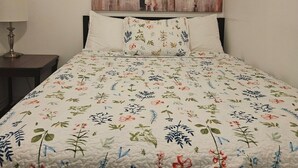 A bedroom with a floral quilted bedspread, a painting of people with umbrellas on the wall, and a bedside lamp.