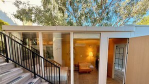 A modern outdoor living space with a glass enclosure, wooden deck, and a staircase leading to an interior room with a sofa and lamp. - Serene Detached Glass Studio—Common Areas with Iconic LA Skyline Views (Los Angeles)