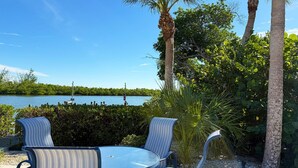 A round table with four chairs, surrounded by lush greenery and palm trees, overlooking a calm body of water. - Sea Oats by the Bay – Pool, Beach & Boat Slips (Englewood)