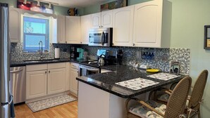 A kitchen with white cabinets, a microwave, and a coffee maker. There is a window with blinds and a rug on the floor. - Sea Oats by the Bay – Pool, Beach & Boat Slips (Englewood)