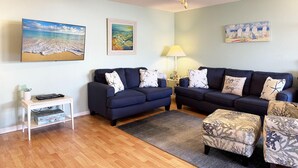A living room with two dark blue sofas, a ceiling fan, and a wooden floor. - Sea Oats by the Bay – Pool, Beach & Boat Slips (Englewood)