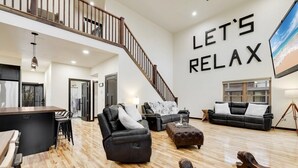 A spacious living area with a wooden floor, a black leather sofa, a black leather armchair, and a wooden coffee table. A staircase leads to an upper level.