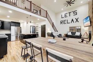 A spacious living area with a wooden dining table, black sofas, and a staircase leading to an upper level.