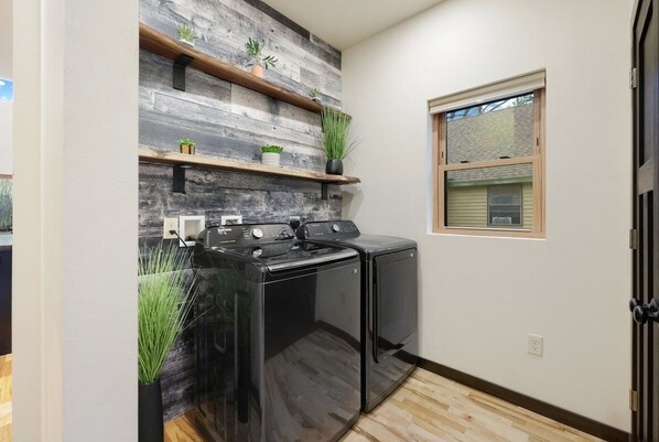 A laundry room with a modern design, featuring a black washer and dryer, wooden shelves with plants, and a window with blinds. - Tubing on the Wolf River - Luxury Experience (New London)
