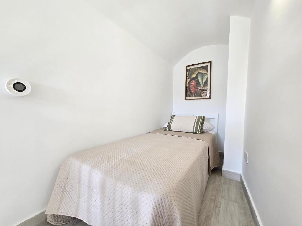 Une petite chambre à coucher, bien rangée, avec un lit simple, une lampe fixée au mur et un tableau encadré.