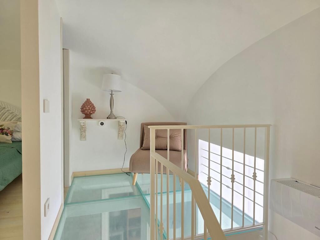 A room with a glass staircase, a bed with a green bedspread, a lamp, and a wall-mounted shelf with decorative items.