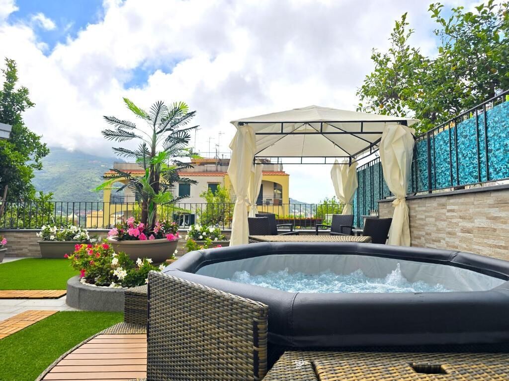 A hot tub with wicker furniture, a covered seating area, and a landscaped garden with flowers and a palm tree.