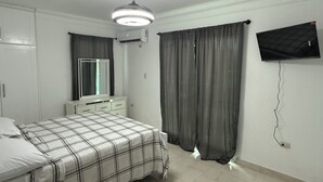 A bedroom with a bed, a dresser, a mirror, a ceiling fan, an air conditioner, a TV mounted on the wall, and a window with curtains. - Modern Getaway with Private Pool, Montecristi (villa vasquez)