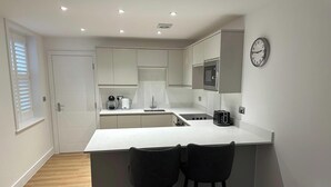 A compact kitchen with a white countertop, two bar stools, a microwave, a toaster, and a clock on the wall. - 59 Whitstable -  One Bedroom Ground Floor Flat (Whitstable)