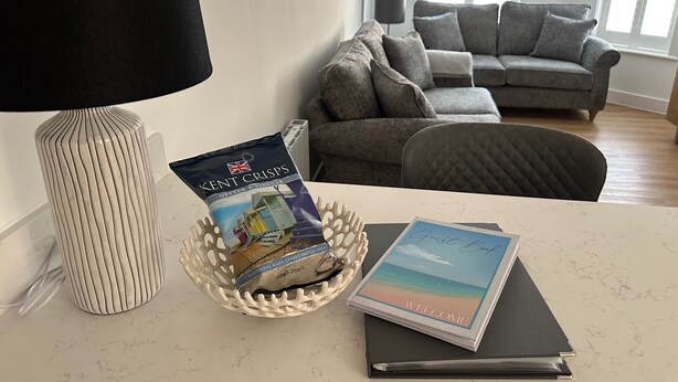 A room with a white lamp, a basket containing a package of Kent Crisps, and a guest book on a table. - 59 Whitstable -  One Bedroom Ground Floor Flat (Whitstable)