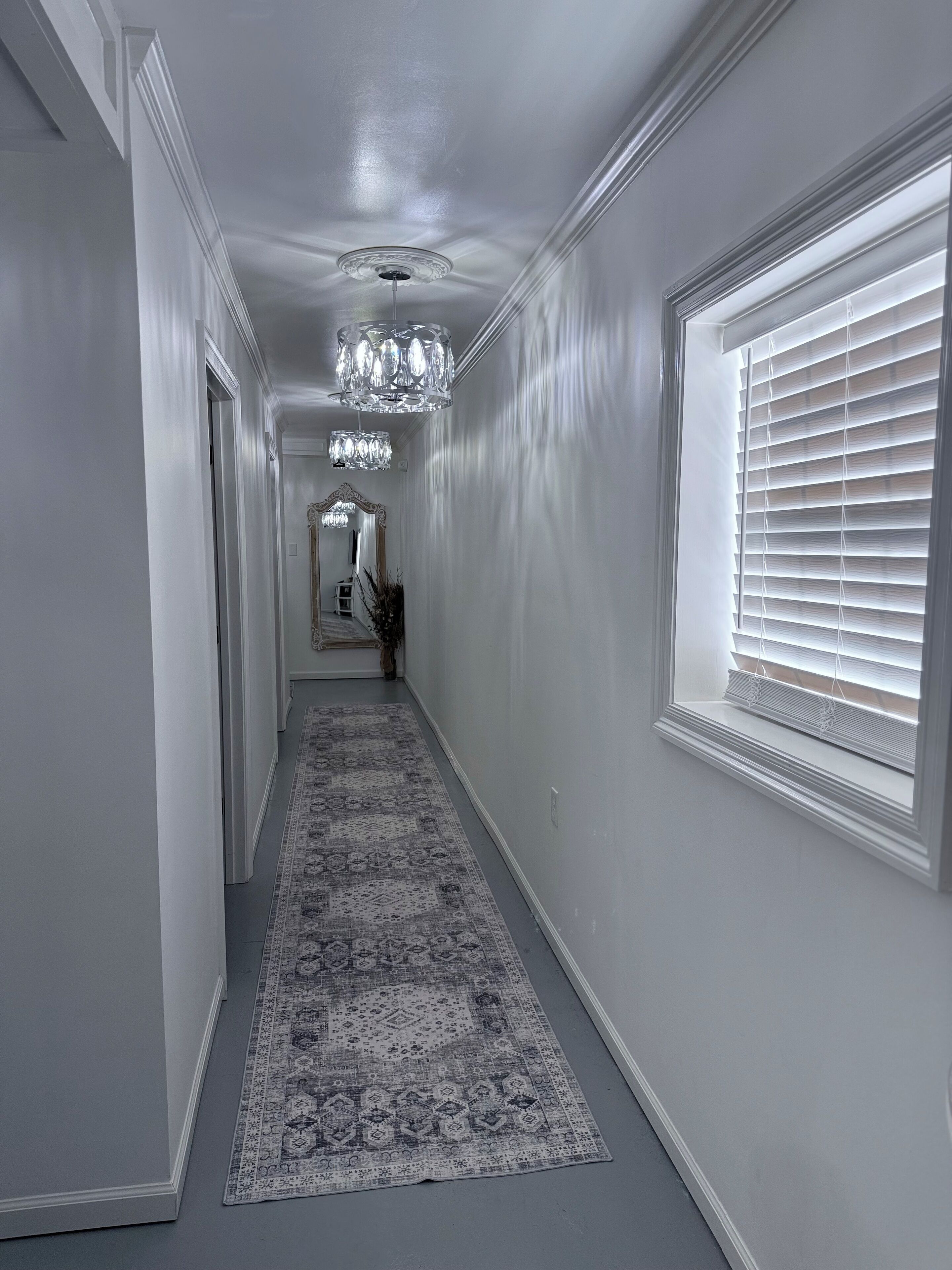 A hallway with a patterned rug, a chandelier, and a window with blinds.
