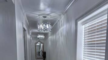 A hallway with a patterned rug, a chandelier, and a window with blinds.