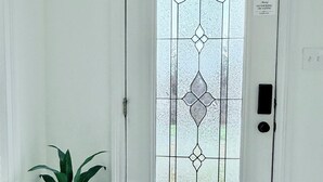 A white door with a frosted glass panel featuring a geometric pattern. A potted plant is placed to the left of the door, and a doormat is in front.