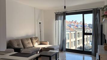 Una sala de estar moderna con un sofá, una mesa de comedor y sillas. Hay un ventanal con cortinas y vistas a edificios.