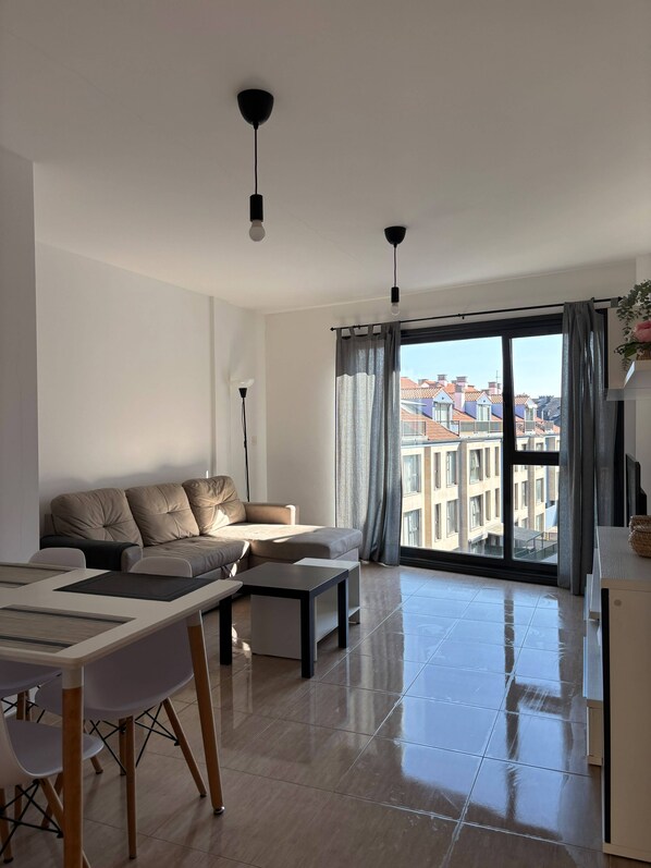 A modern living room with a sofa, a dining table, and chairs. There is a large window with curtains, and a view of buildings outside.