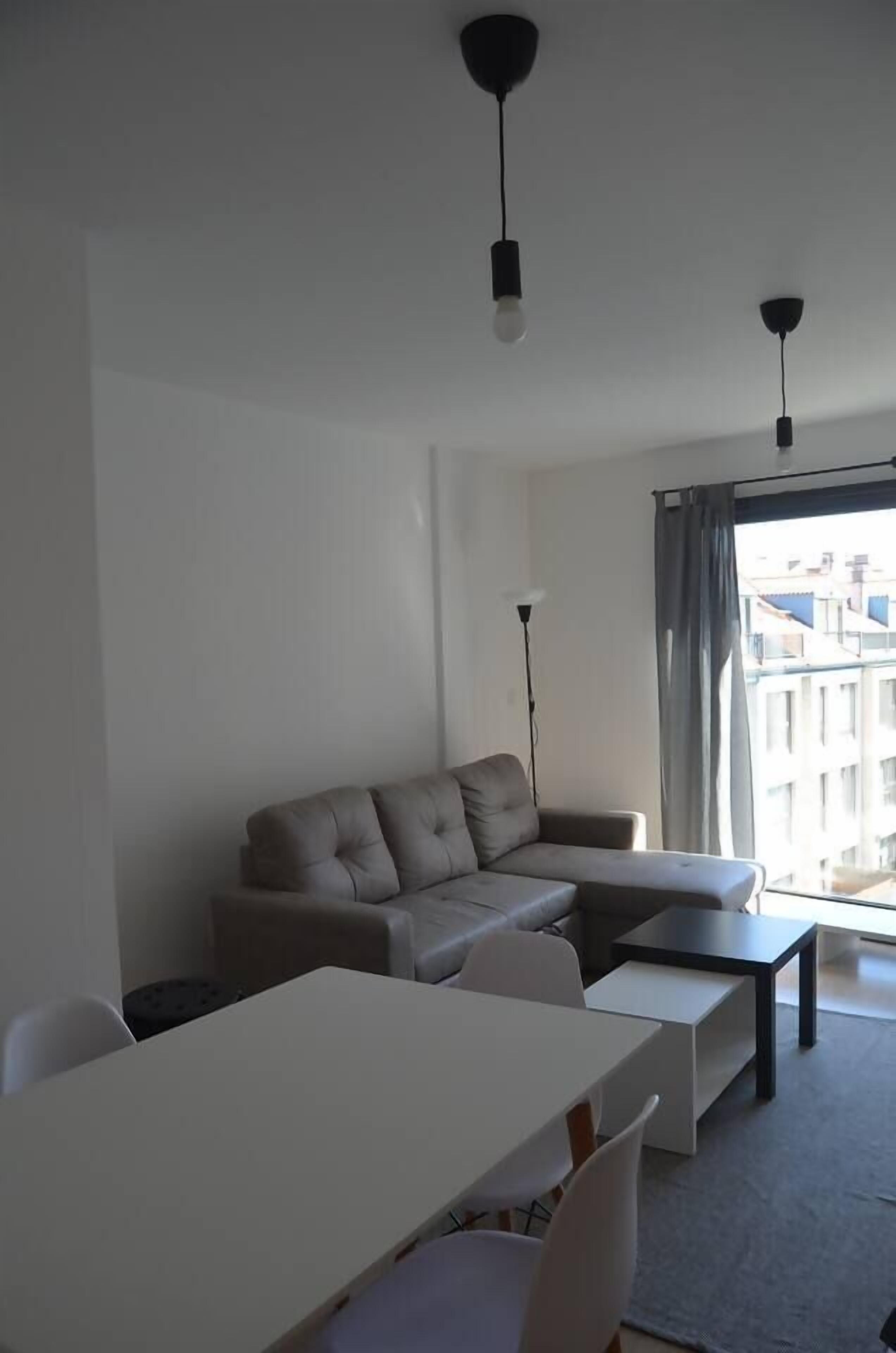 Una sala de estar moderna con un sofá, una mesa de comedor y sillas. Hay dos lámparas colgantes y una lámpara de pie. La habitación tiene un ventanal con vistas a edificios.