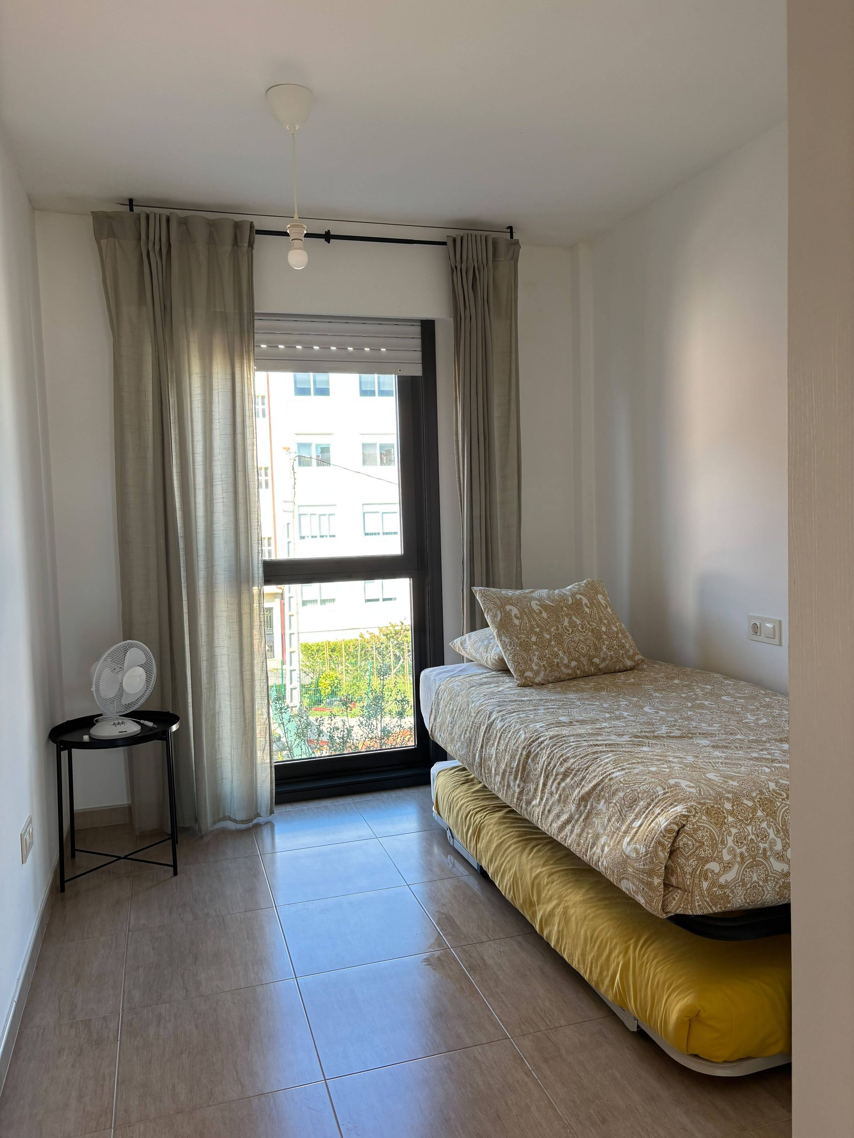 Un dormitorio con cama, ventilador y vista a edificios a través de una ventana.