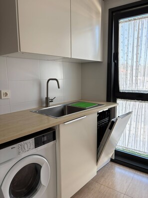 A compact kitchen with a built-in washing machine, a sink, and a window with a patterned curtain. - Lovely apartment with garage in Ares (Ares)